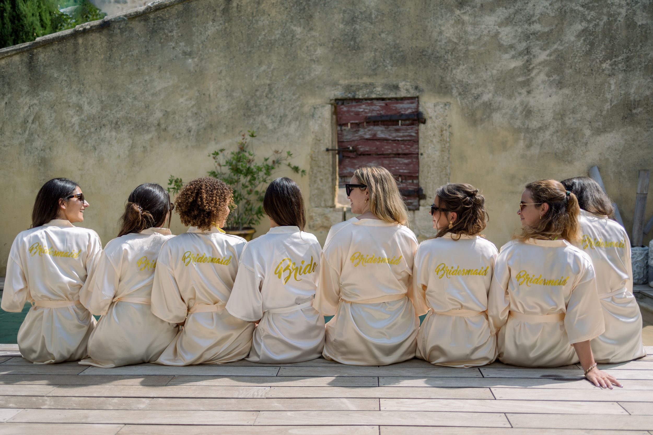 Wedding party members sit together in matching robes against a rustic stone wall.