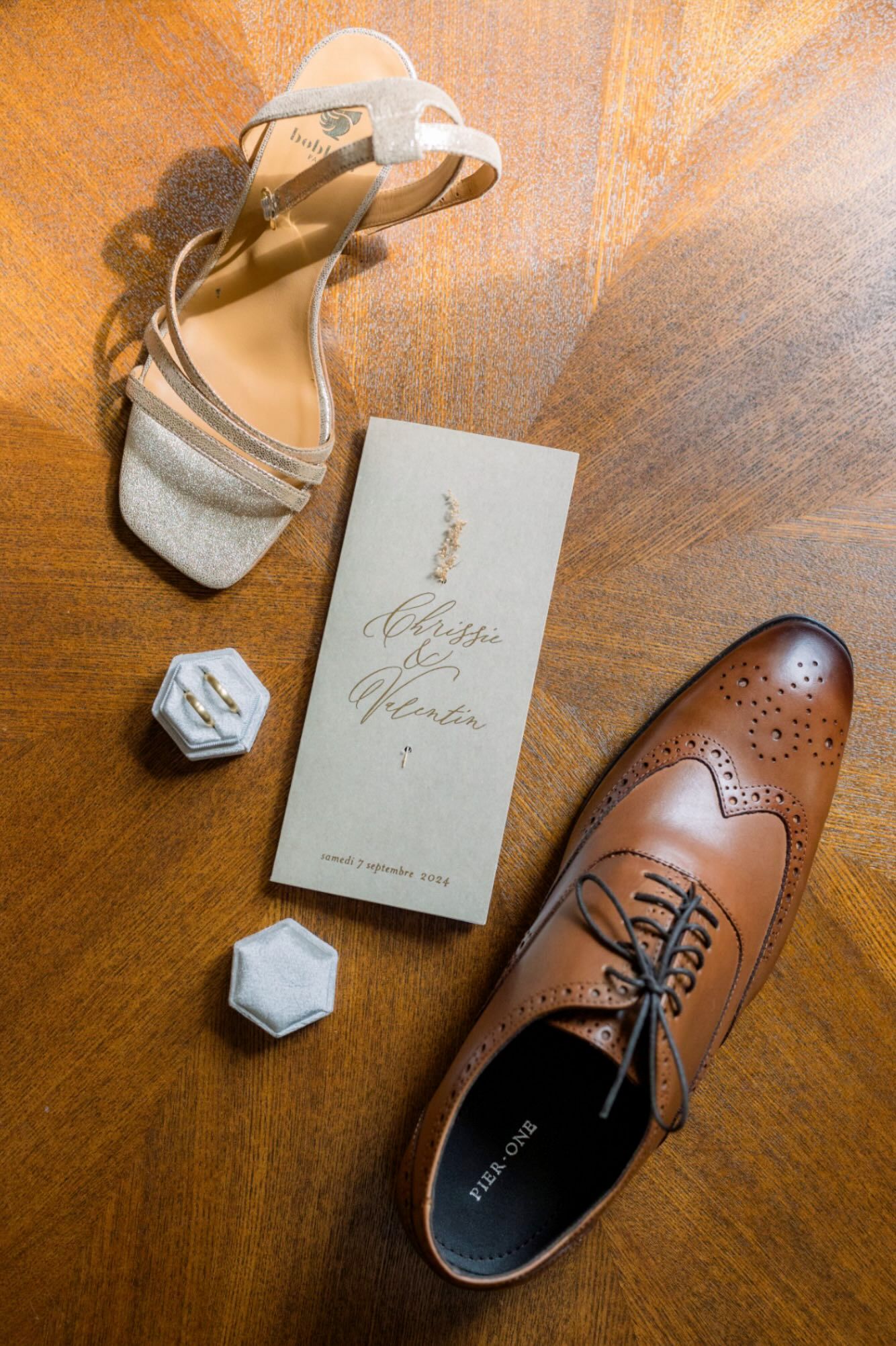 Wedding accessories including a brown leather dress shoe, silver sandal, hexagonal ring boxes and invitation on wooden surface.