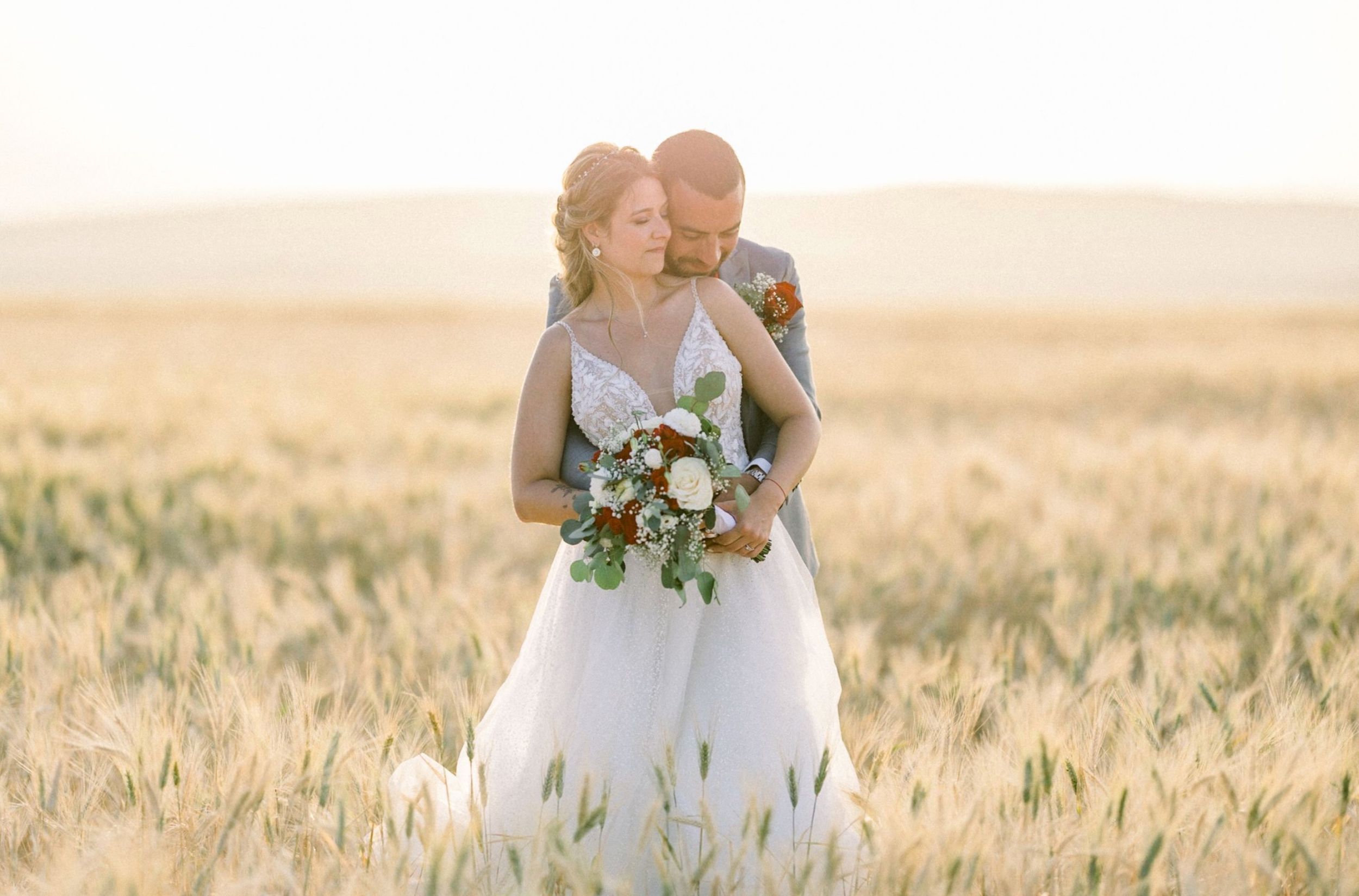 A bride in a white wedding dress holds a bouquet while standing in a golden wheat field at sunset.