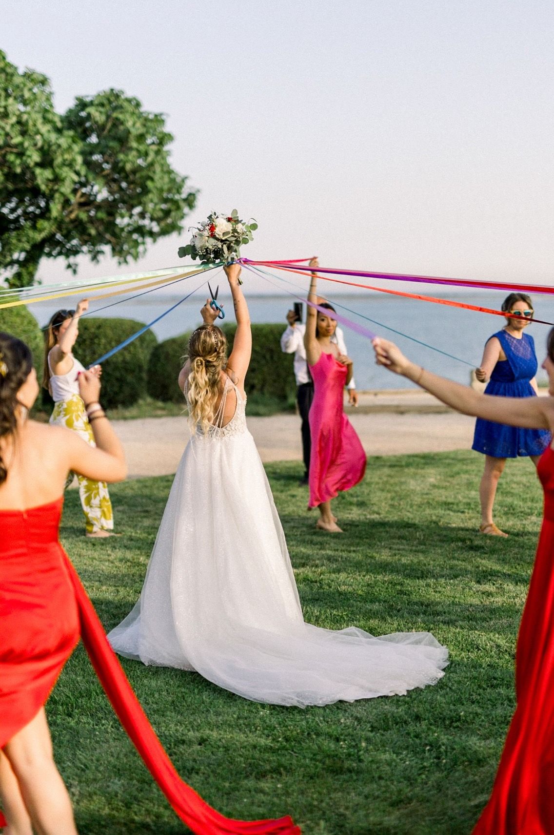 A wedding party dances and twirls on a grassy hilltop at sunset wearing red and blue dresses with a flowing white gown.