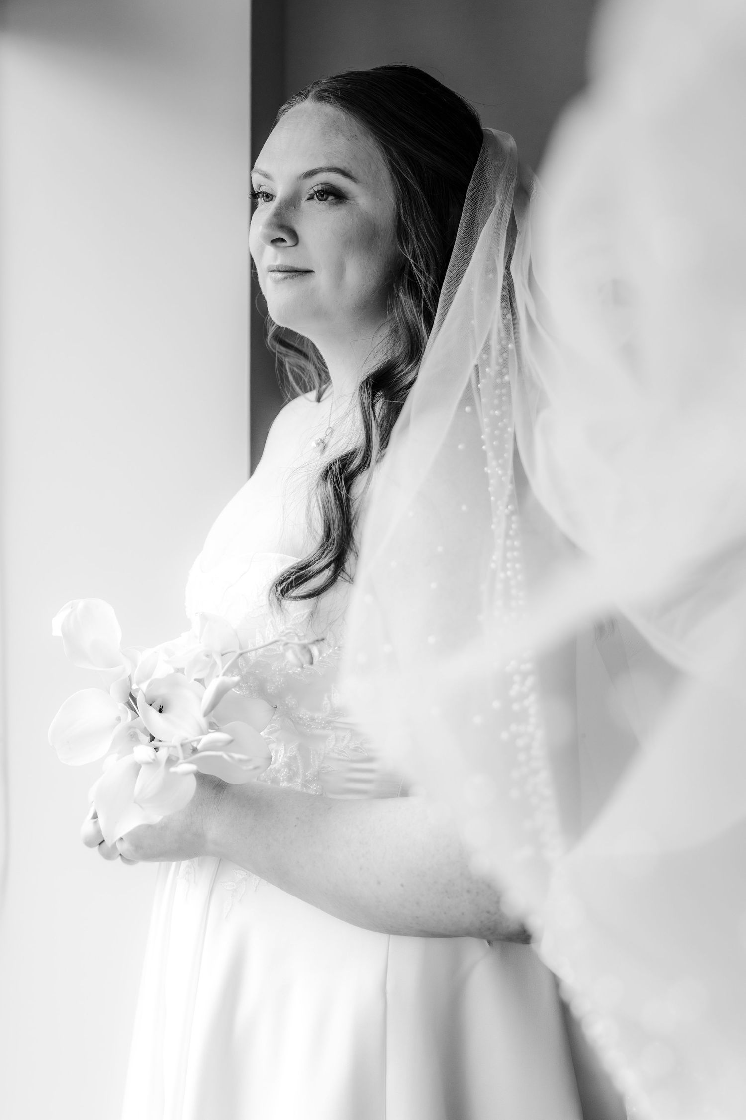 Bride in white gown with flowing veil stands before mirror in black and white bridal portrait.