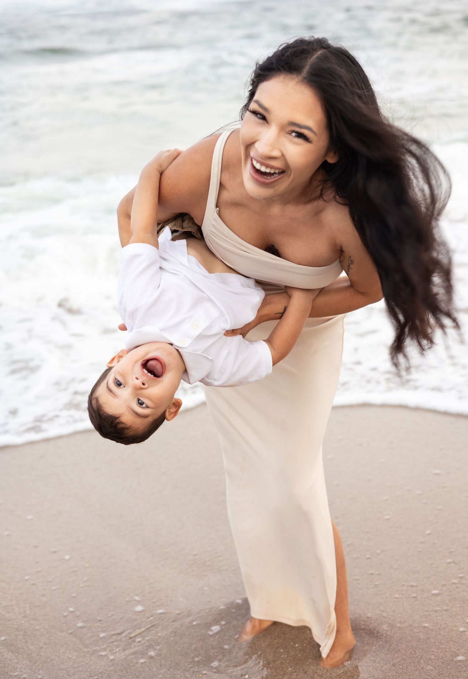 A joyful moment captured at the beach as a parent playfully holds a laughing child upside down near the ocean waves.