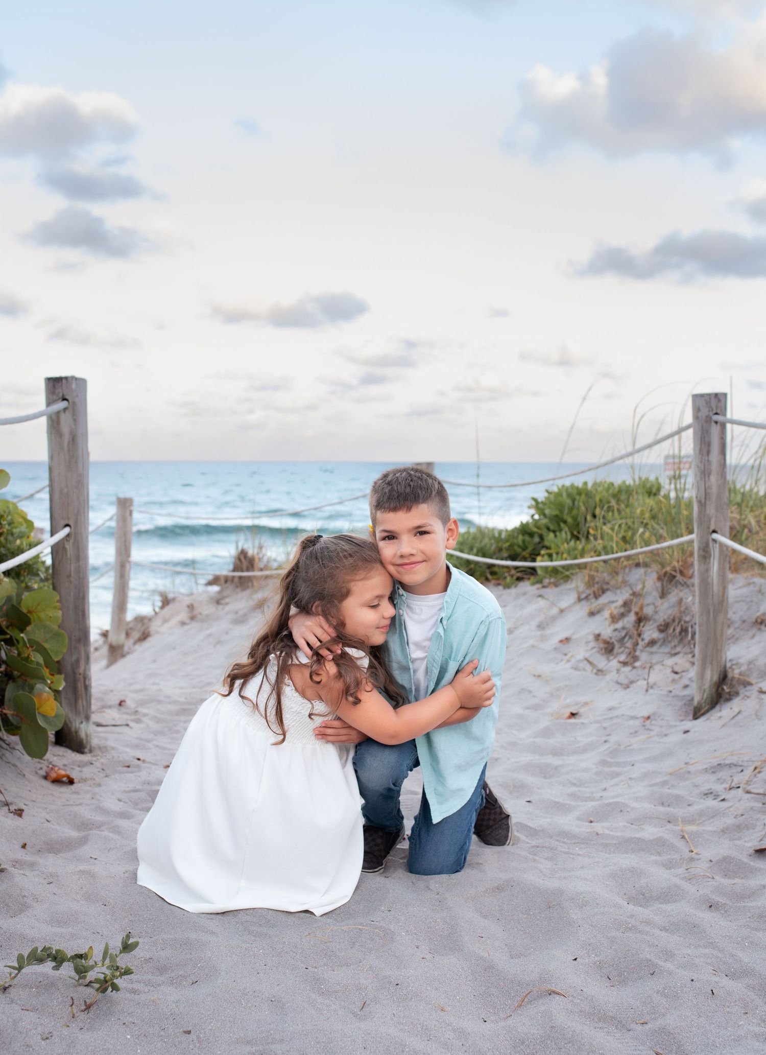 Family shares an intimate moment together on a sandy beach path at sunset with ocean waves and cloudy sky in background.