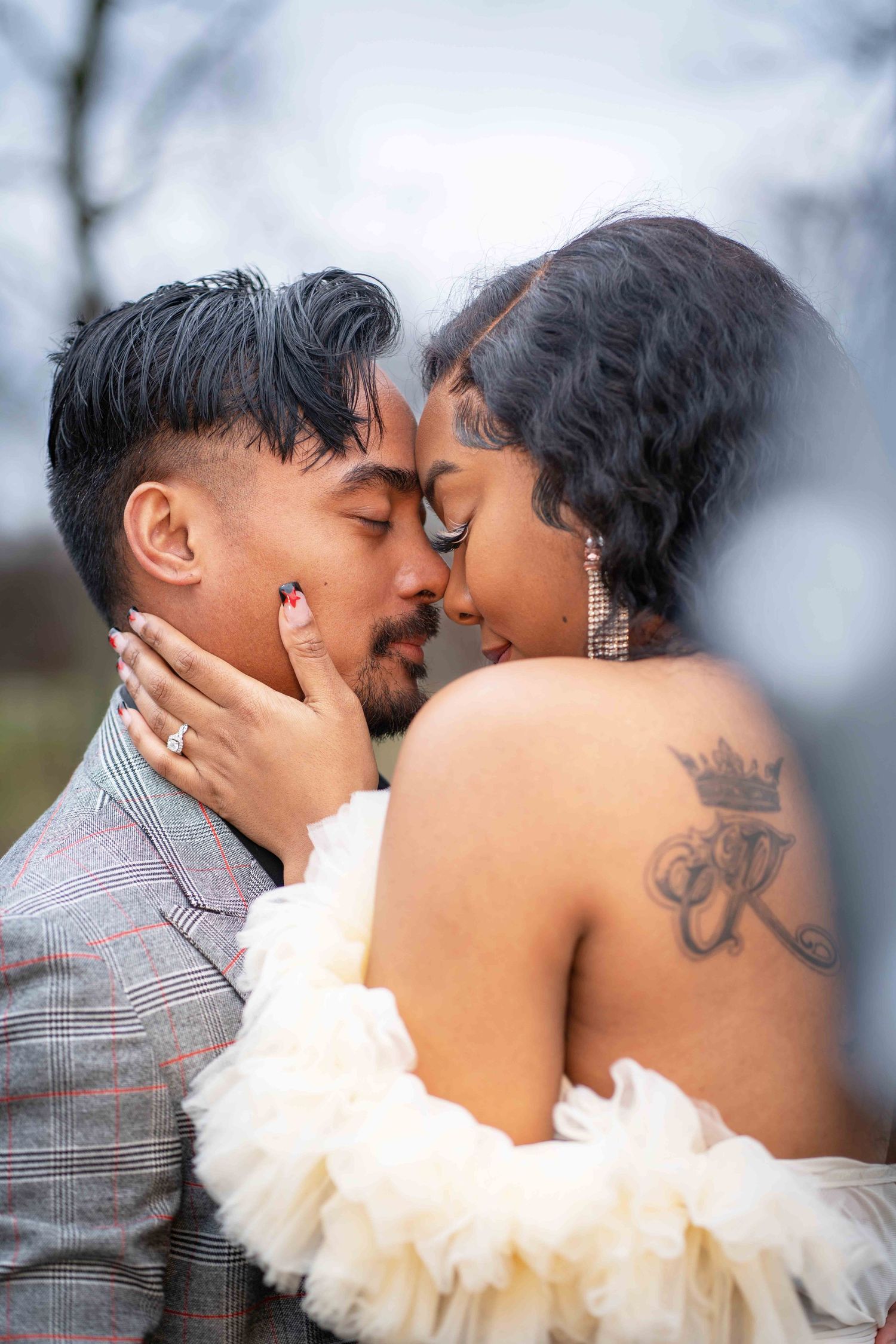 A romantic couple shares an intimate moment outdoors, with one wearing a plaid suit and the other in an off-shoulder white dress.