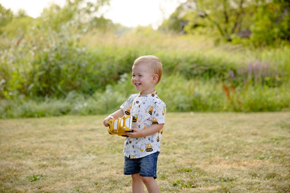 Young child in star-printed shirt and denim shorts plays in sunny meadow with water bottle on summer day.