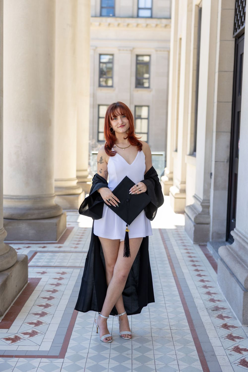 Young woman in white dress and black graduation gown holding cap, posing between columns of university building.