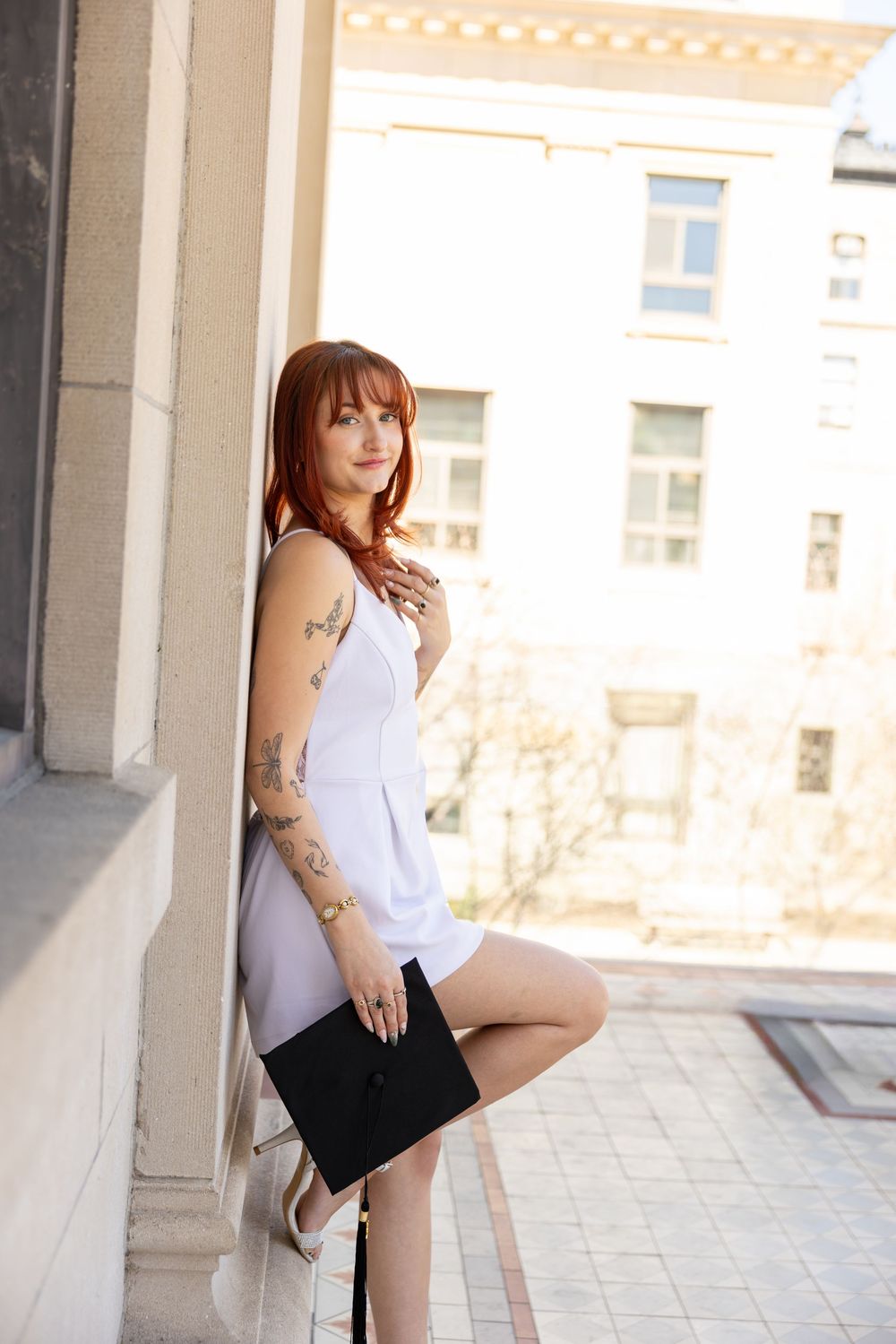 Young redhead woman with tattoos in white dress holds black clutch, leaning against stone column outdoors.