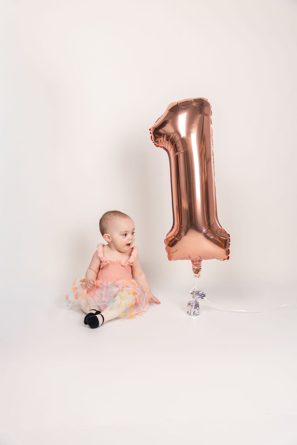 Baby in pink dress sits beside large rose gold number 1 balloon against white backdrop for first birthday photoshoot.
