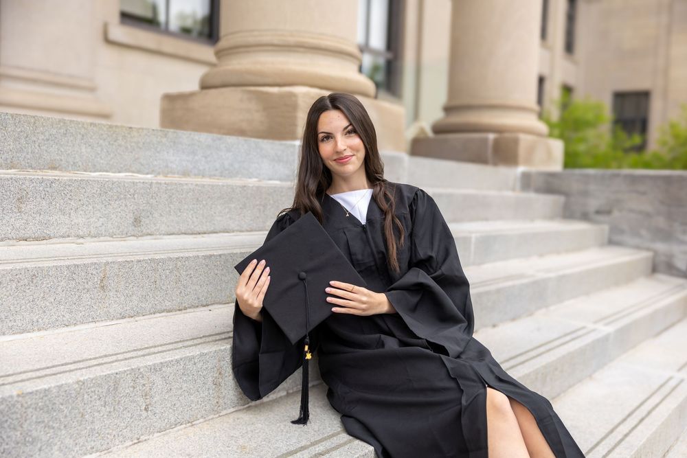 Young woman in black graduation gown and cap sitting on stone steps outside university building.