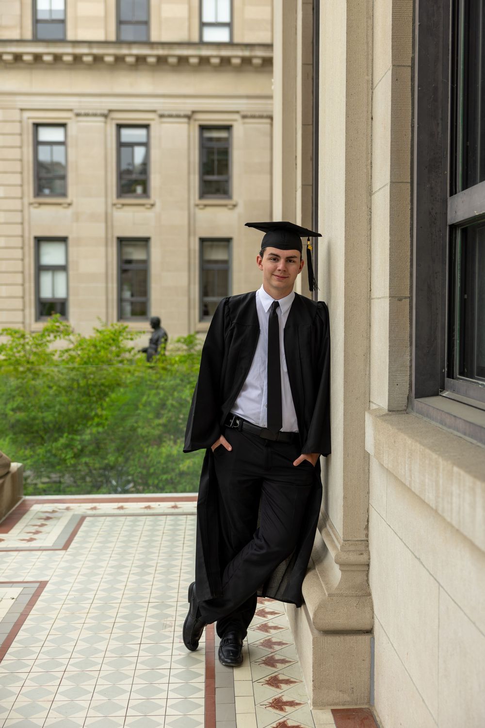 Young male graduate in black cap and gown leans against stone column on university campus balcony.