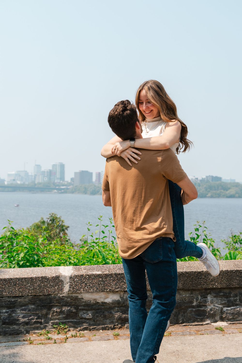A couple embraces on a waterfront overlook with a city skyline visible across the water on a sunny day.