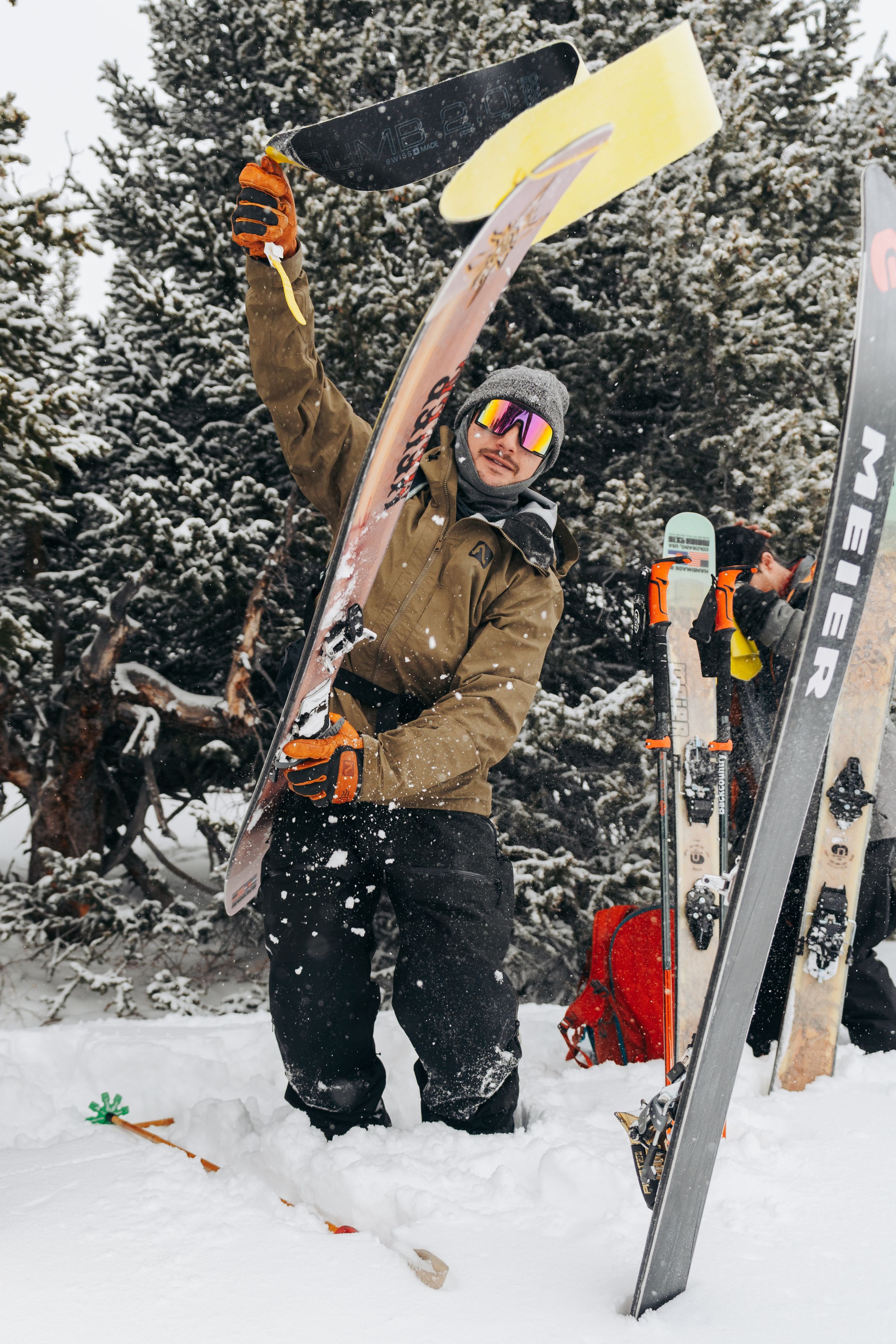 Skier in brown jacket raises ski gear triumphantly against snowy evergreen forest backdrop on winter mountain slope.