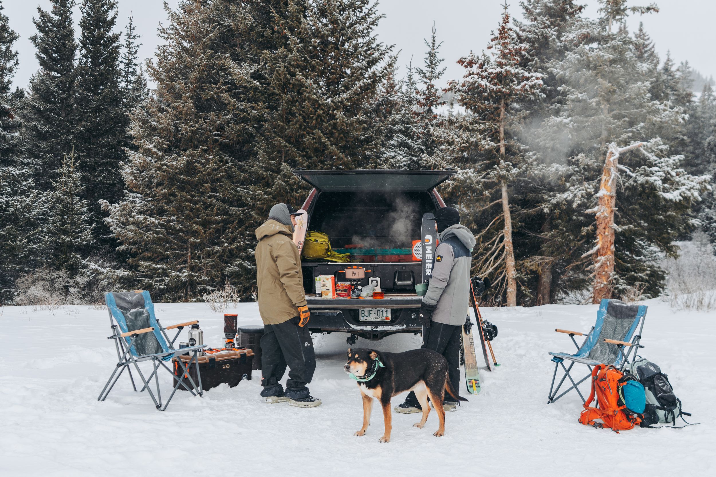 A group gathers around camping chairs and equipment in a snowy winter forest setting for outdoor recreation.