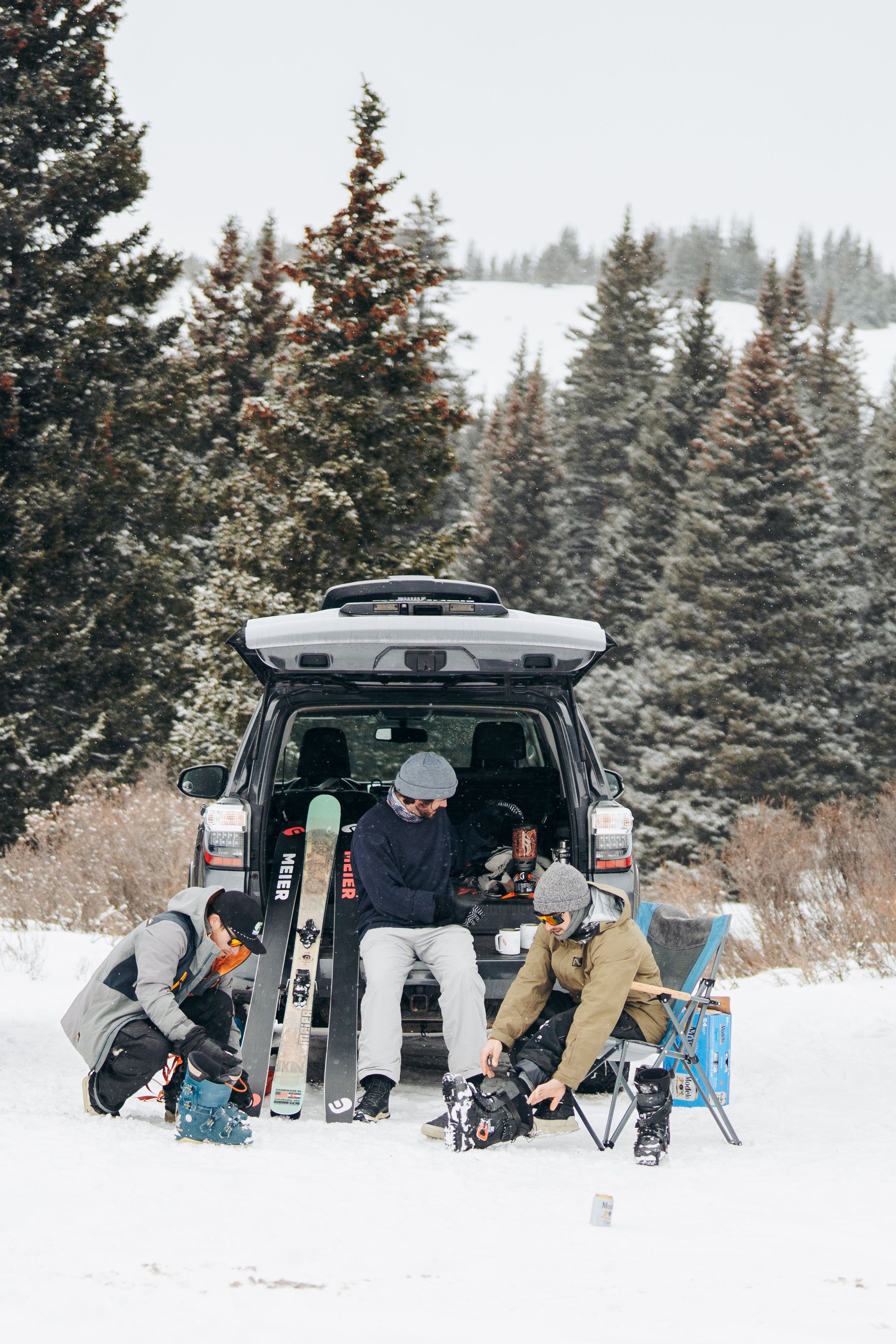 Winter sports enthusiasts gather around SUV with roof cargo box in snowy mountain setting surrounded by evergreens.