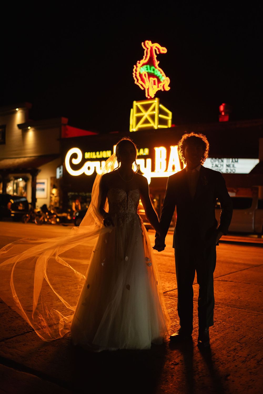 Silhouettes of a couple in formal attire stand against dramatic nighttime lighting with neon signs.