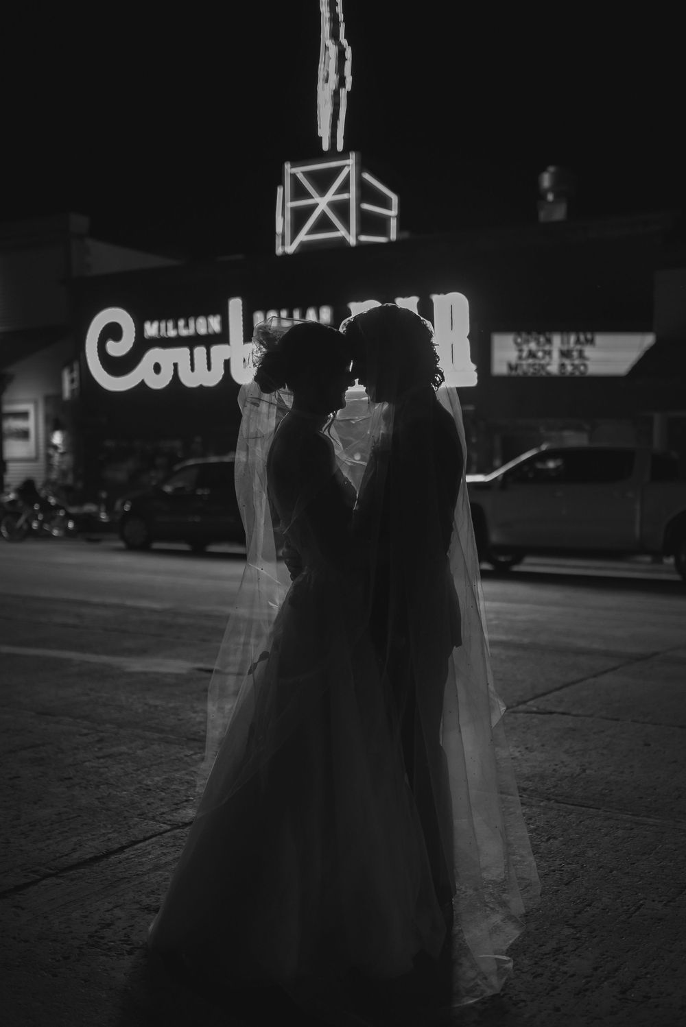 Silhouettes of people walking at night on a dimly lit street with illuminated storefronts in the background.