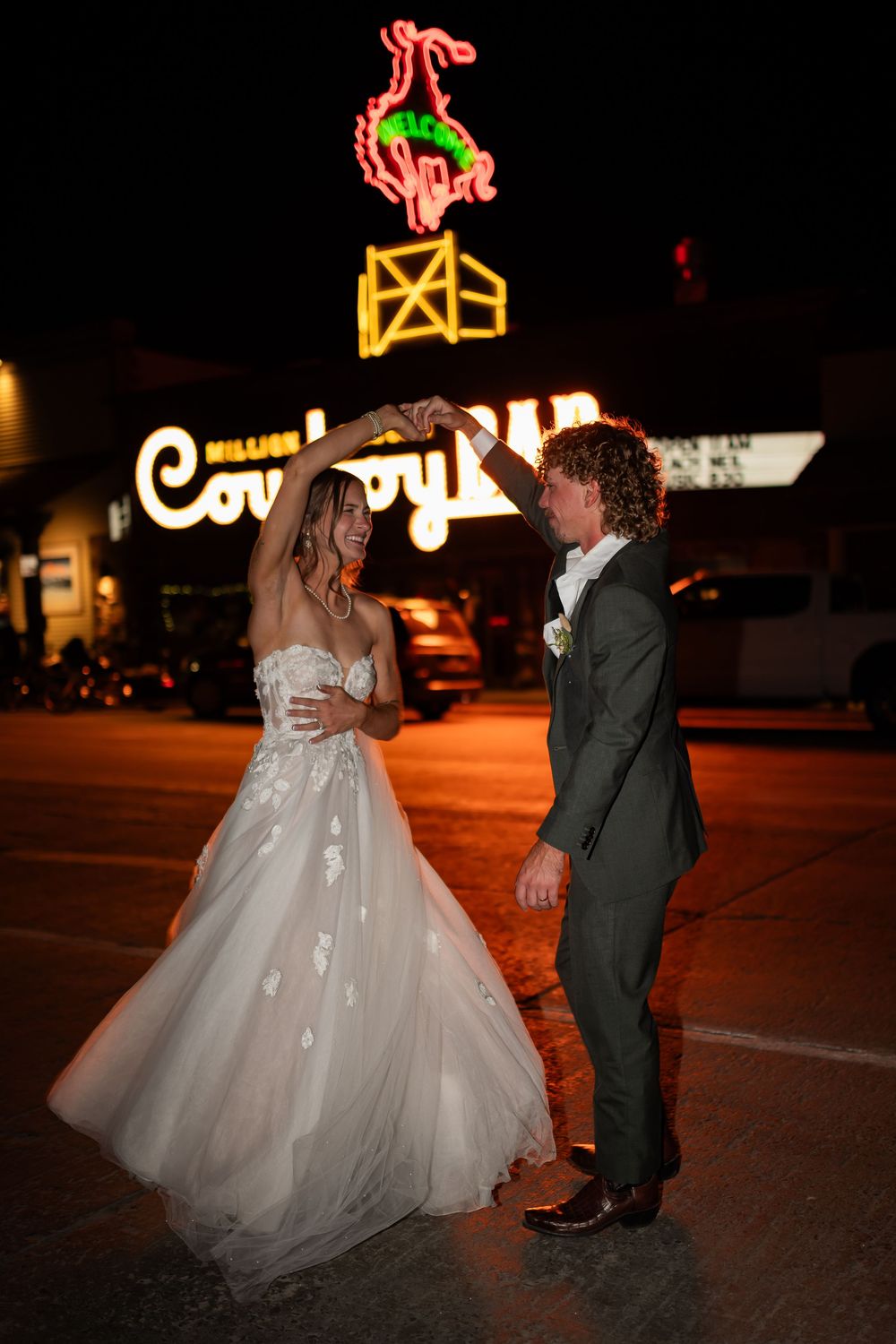 A couple in formal wedding attire dances together at night with illuminated neon signs glowing in the background.