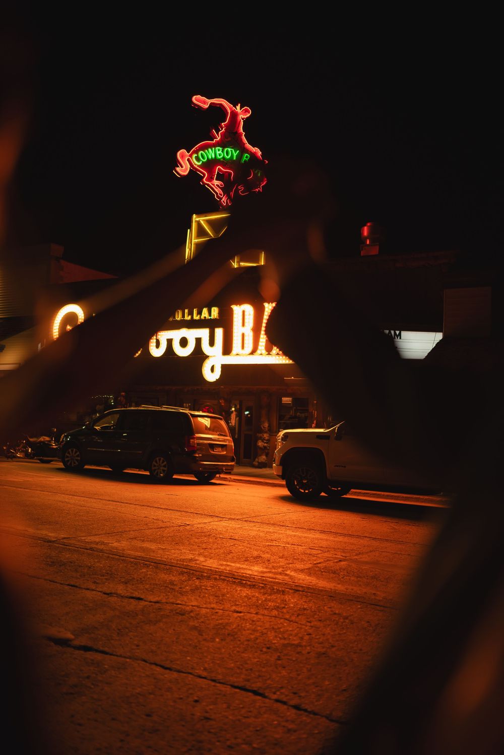 Vintage neon signs illuminate parked cars on a dark street at night creating a moody retro atmosphere.