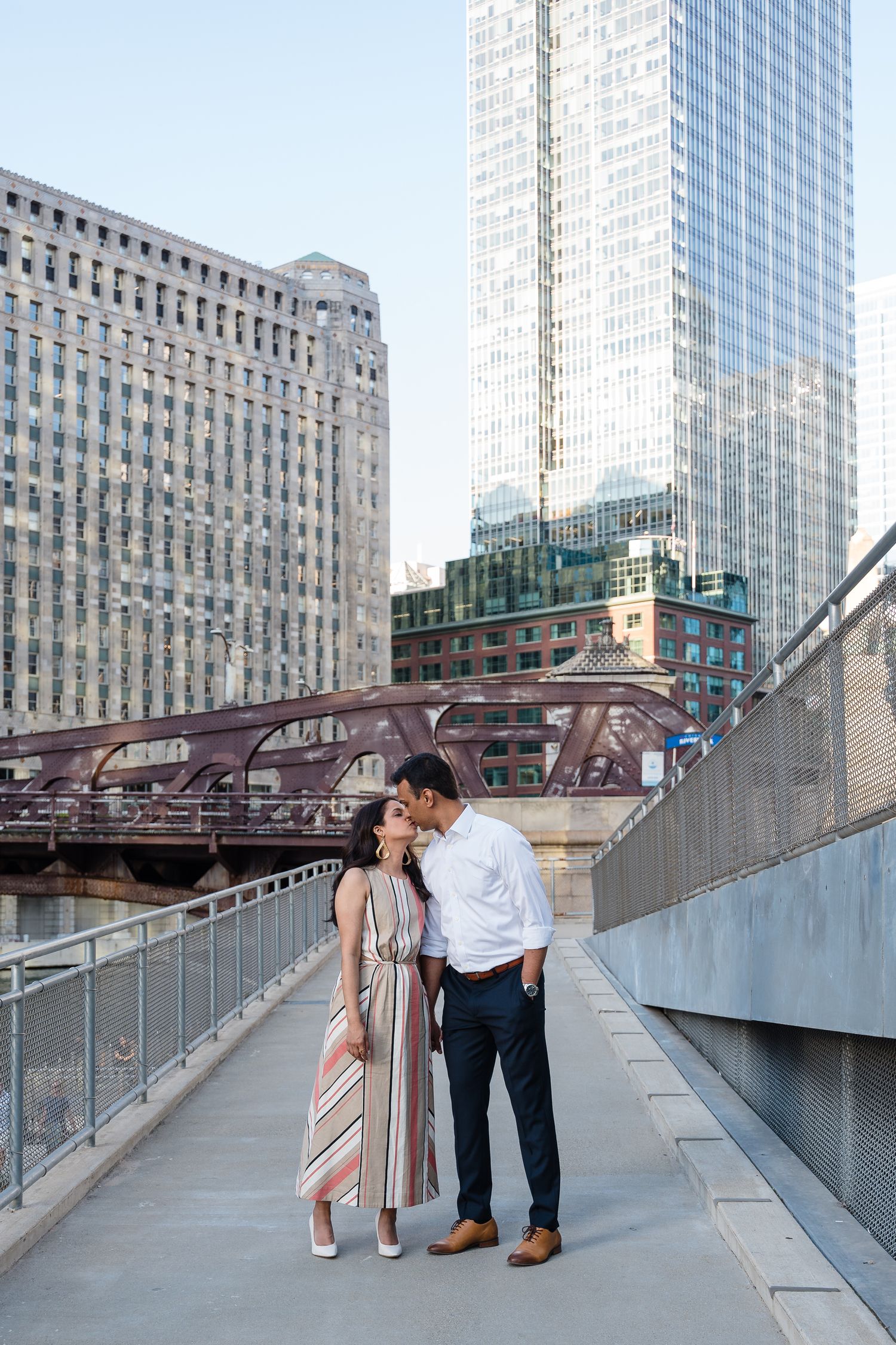 A couple shares a romantic moment on a pedestrian bridge in downtown Chicago with urban architecture in the background.