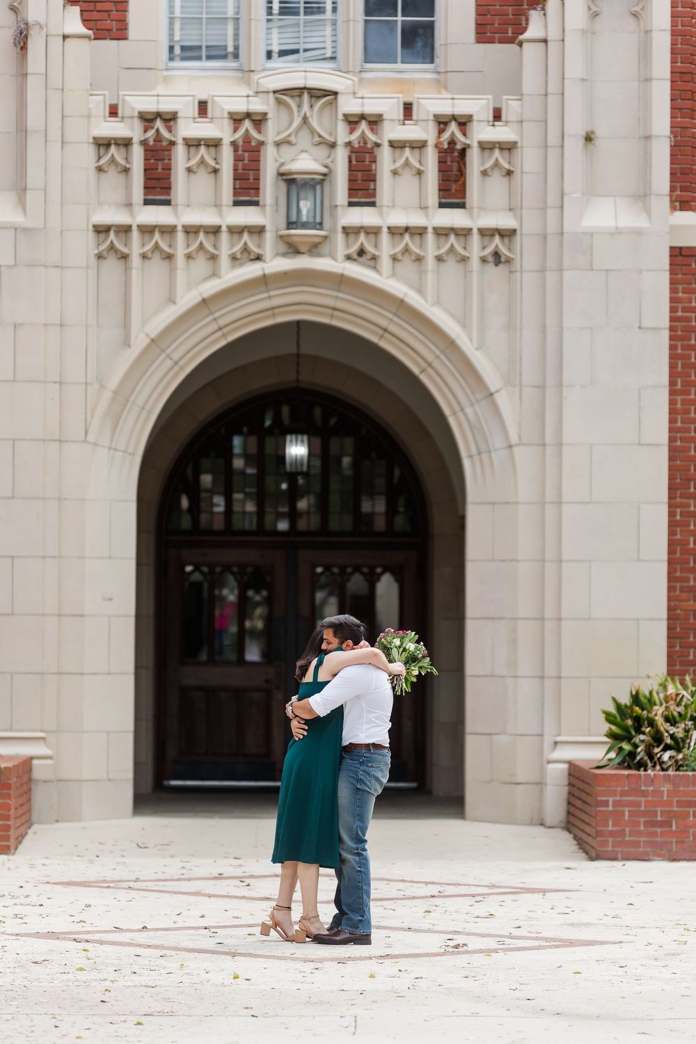 A romantic couple embraces in front of an ornate brick and stone building with arched entrances.