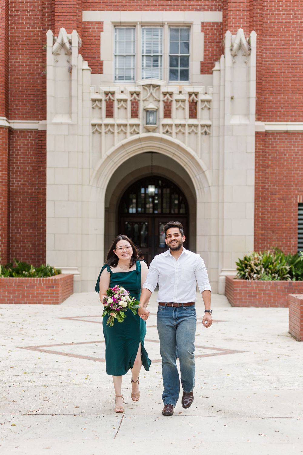A couple runs joyfully out of a grand brick university building with Gothic architectural details.