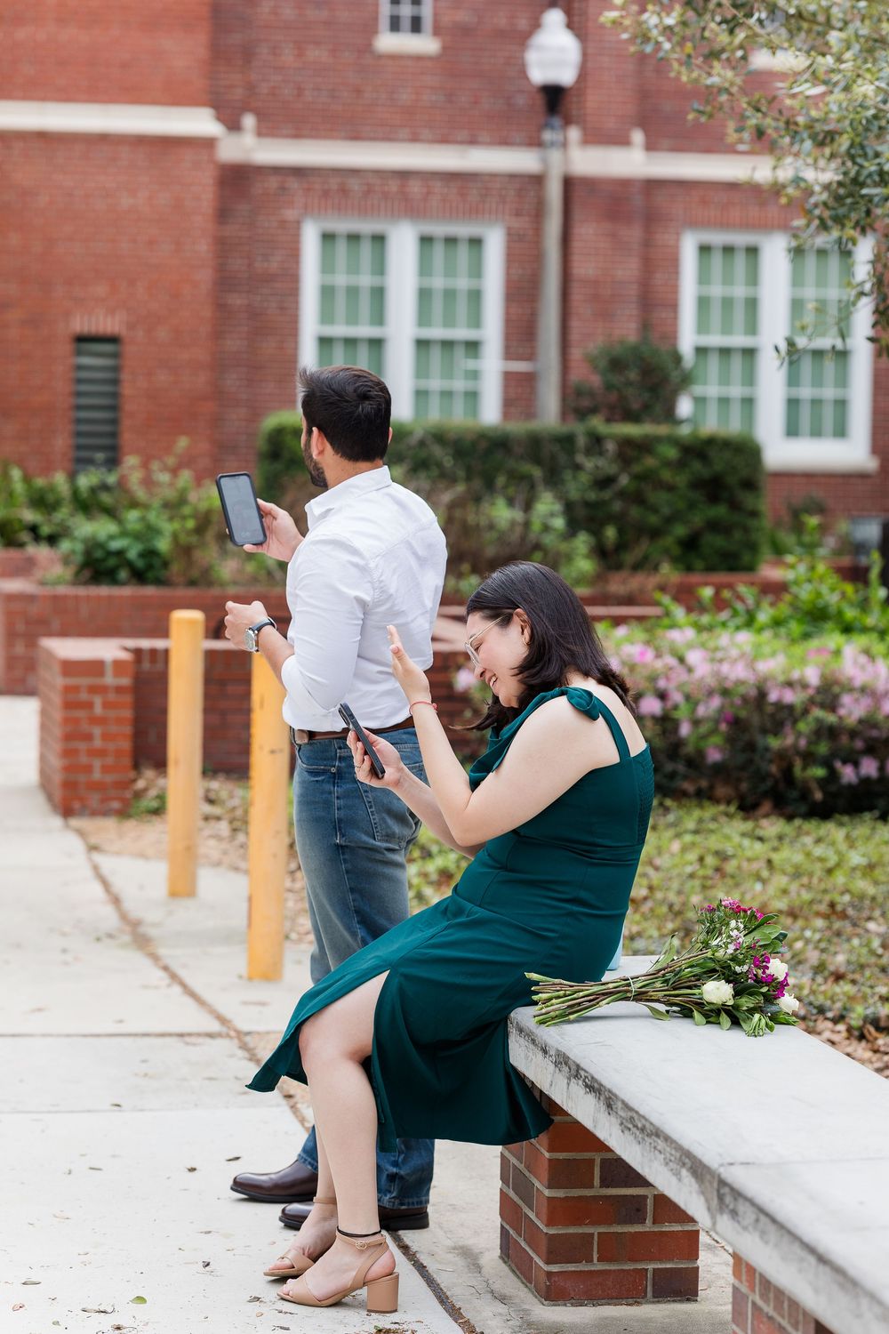 Two people checking their phones while standing and sitting near a brick building on a sunny day.