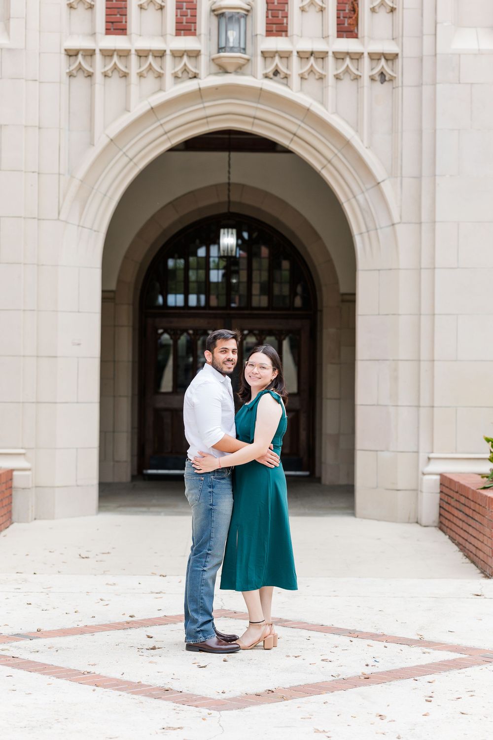 Couple embracing in front of an ornate stone archway entrance to a classic brick building.