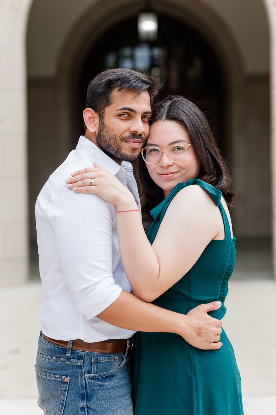 A couple in a green dress and white shirt embrace lovingly in an outdoor setting with arched columns.