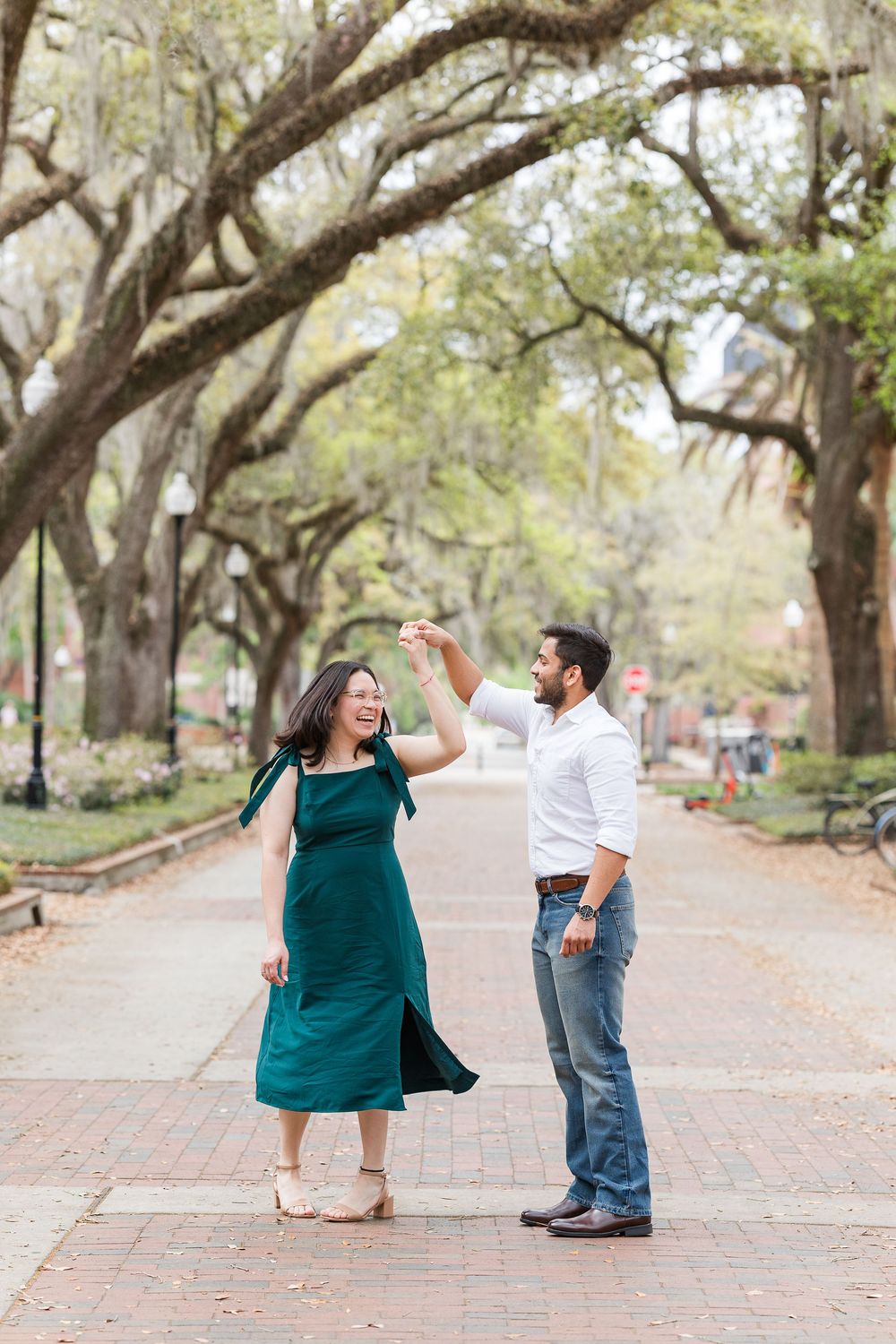A couple dancing and twirling under moss-draped oak trees on a brick pathway.