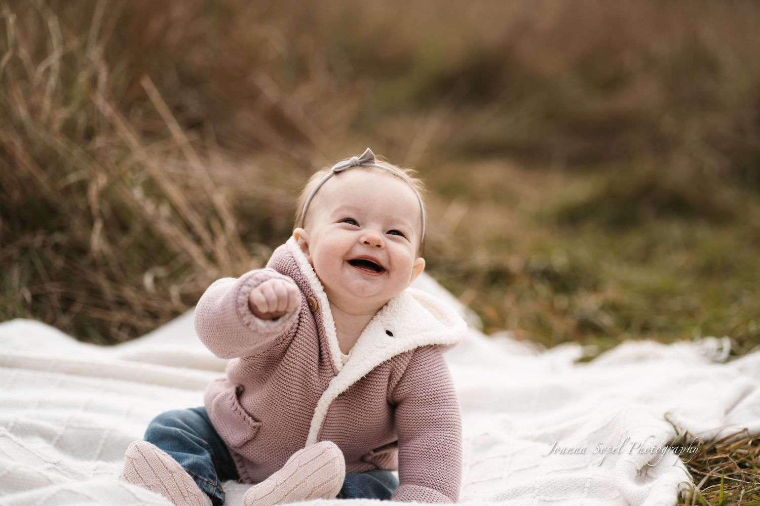 A baby in pink clothing lies on a white blanket outdoors while smiling and playing during a photo session.