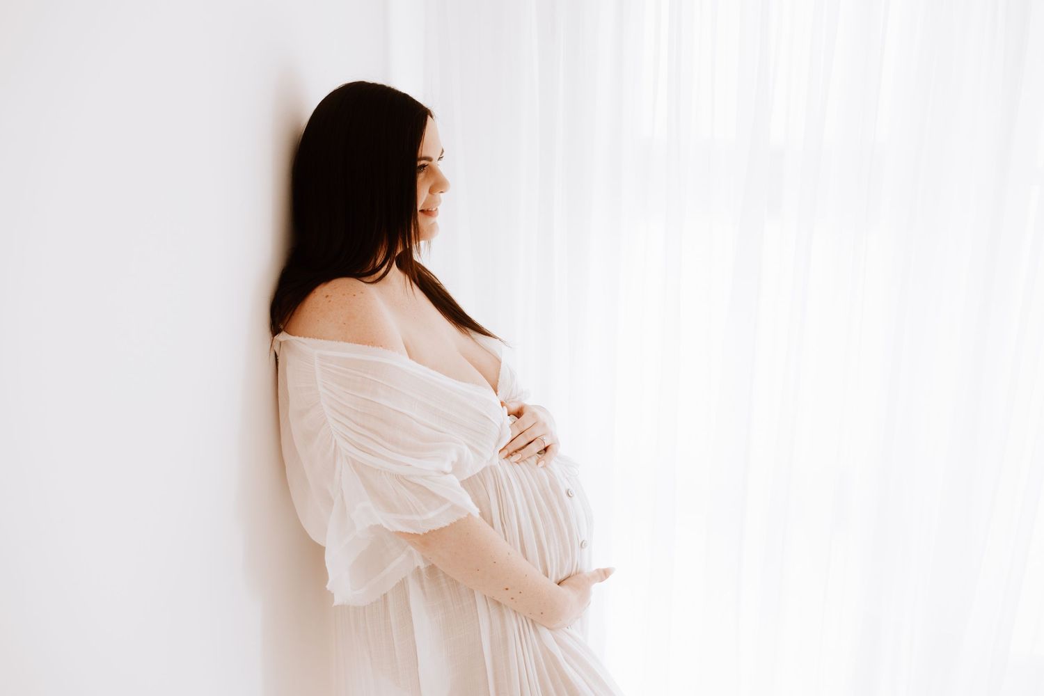 Side view of pregnant woman in white dress against bright window.