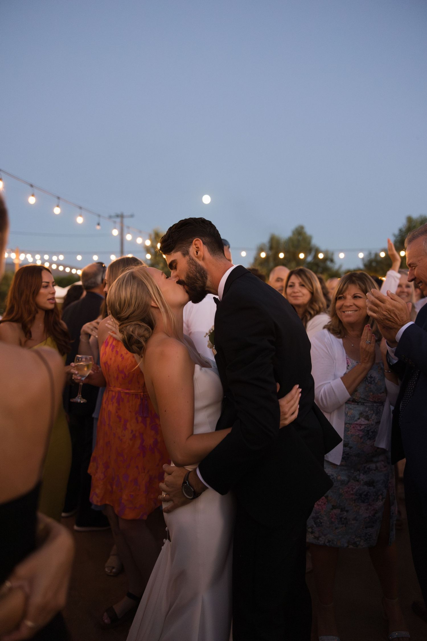 Bride and groom kissing on the dance floor at dusk surrounded by guests and twinkle lights at Tuscan Rose Ranch in Santa Paula, California