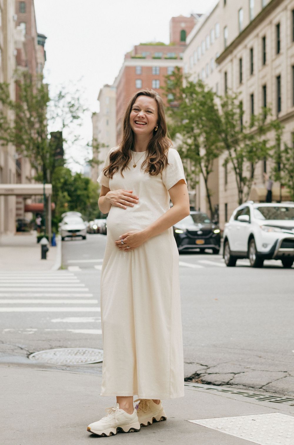 A person in a flowing cream-colored dress stands on a city street corner with urban buildings in the background.