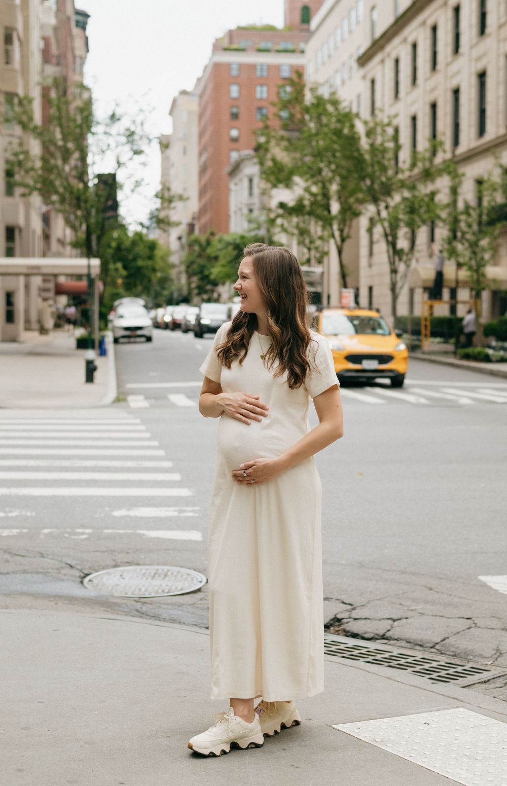 A pregnant woman in a flowing white dress stands on a New York City street corner with yellow taxis visible in the background.