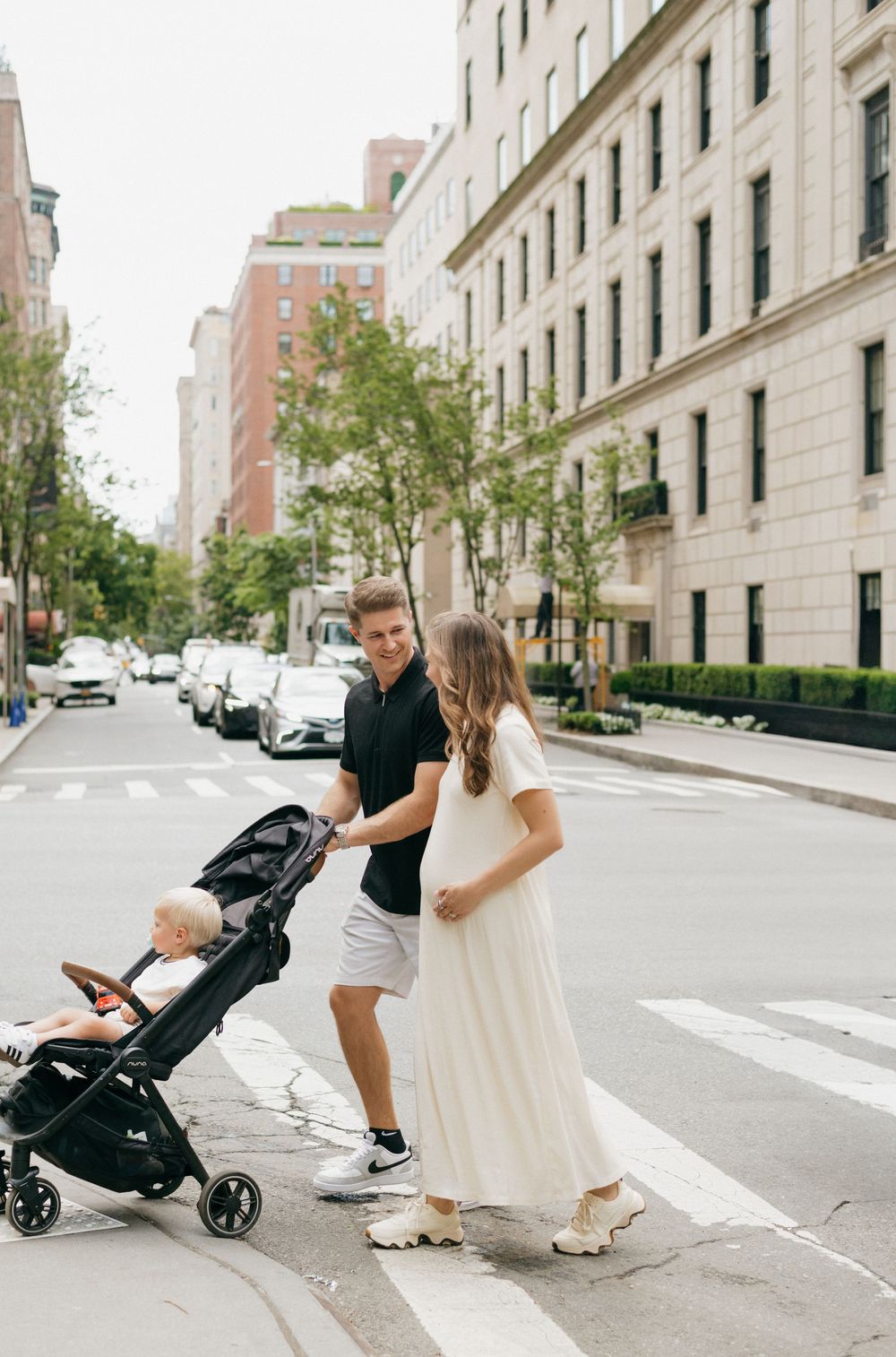 A family walks down a city street with a stroller while wearing casual attire on a sunny day.