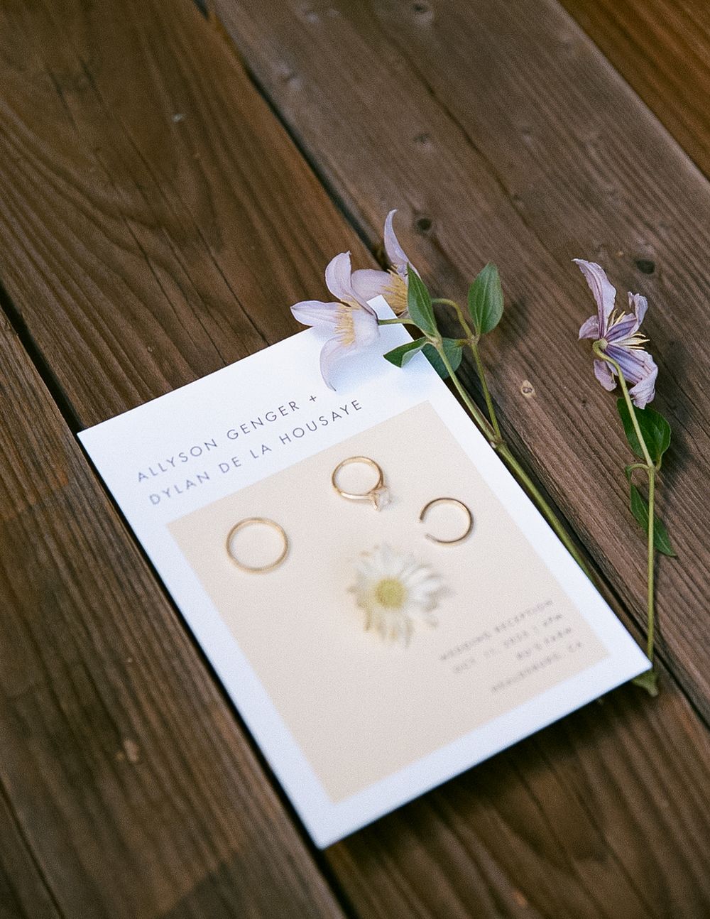 Wedding invitation with gold rings and delicate purple flowers arranged on a rustic wooden surface.