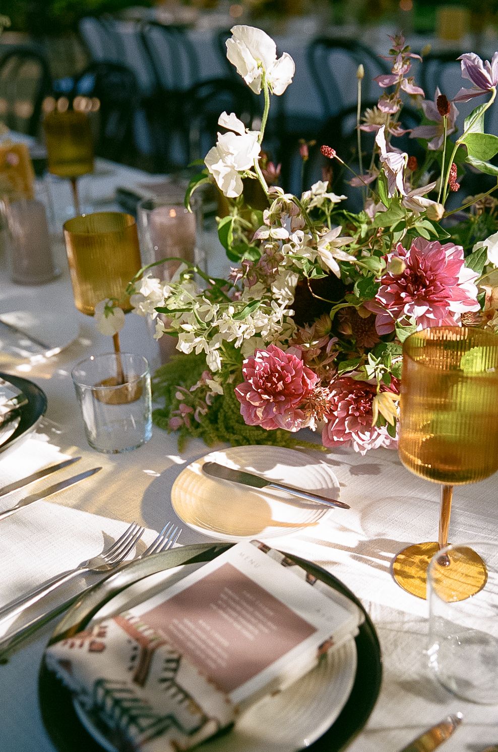 Elegant table setting with pink carnations and white flowers in centerpiece, amber glassware, and place settings at golden hour.