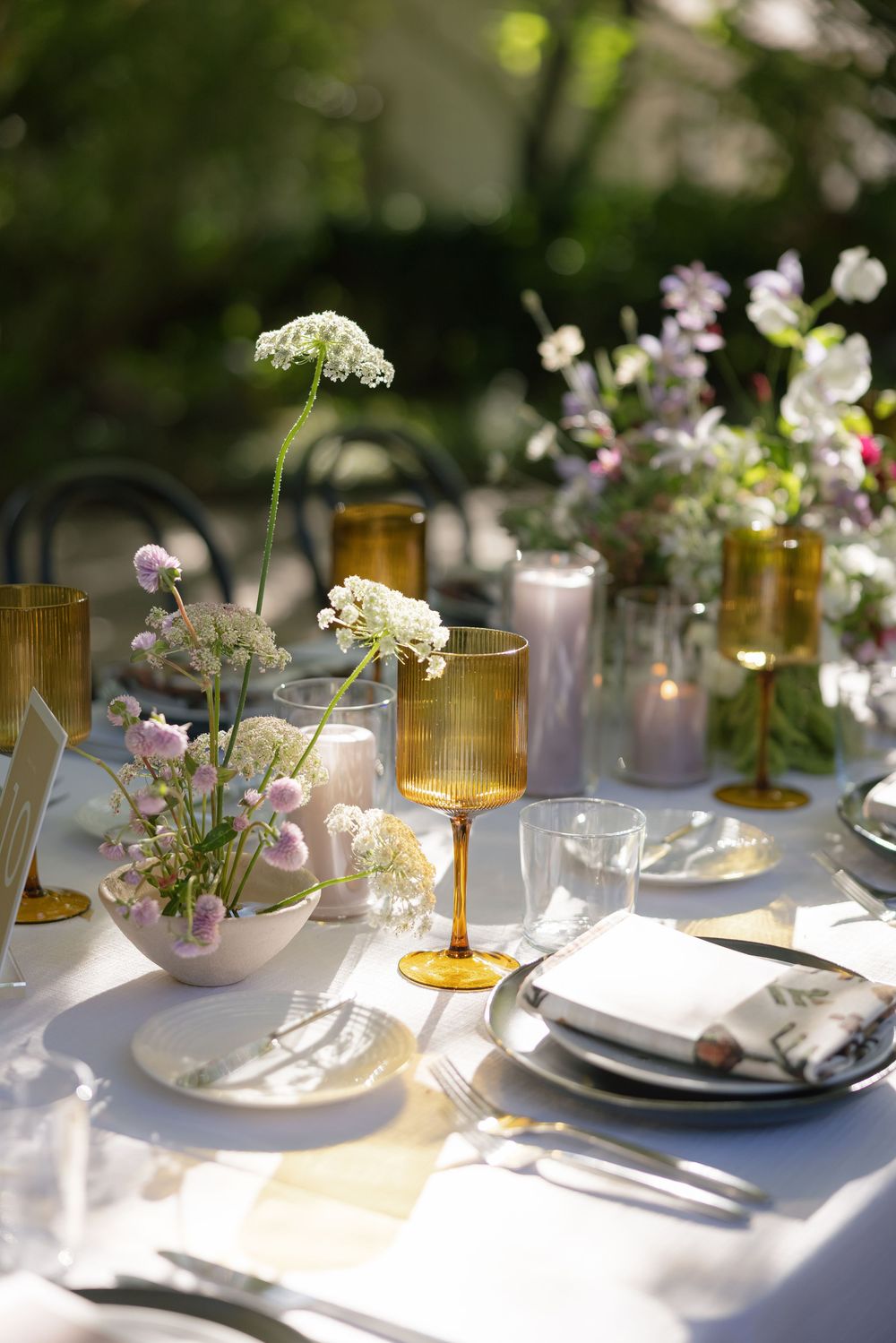 Elegant outdoor table setting with amber glassware and delicate floral arrangements in soft natural lighting.