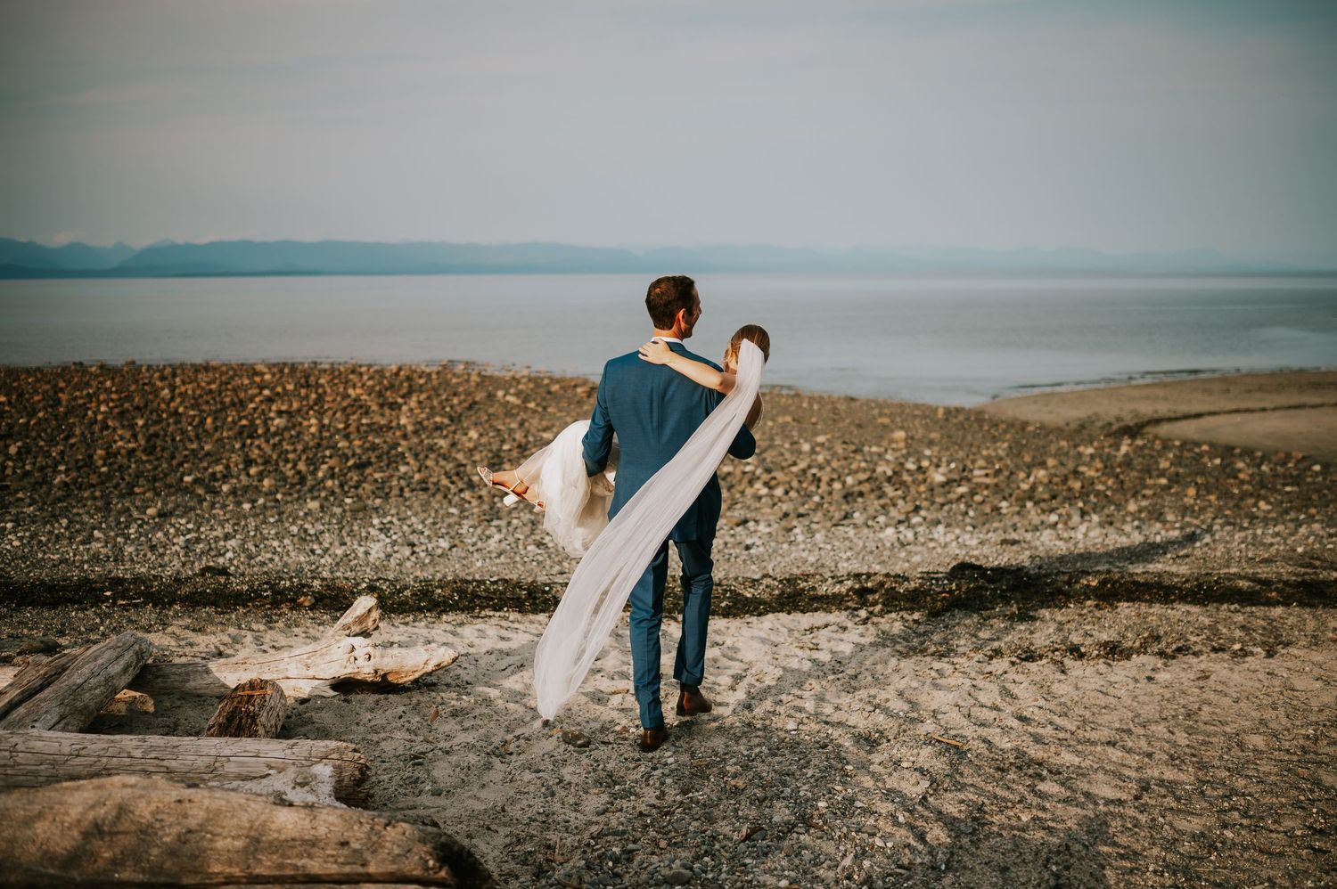 Wedding photography of couple on Williams Beach, groom carrying bride during golden hour with ocean, sand and mountains in background