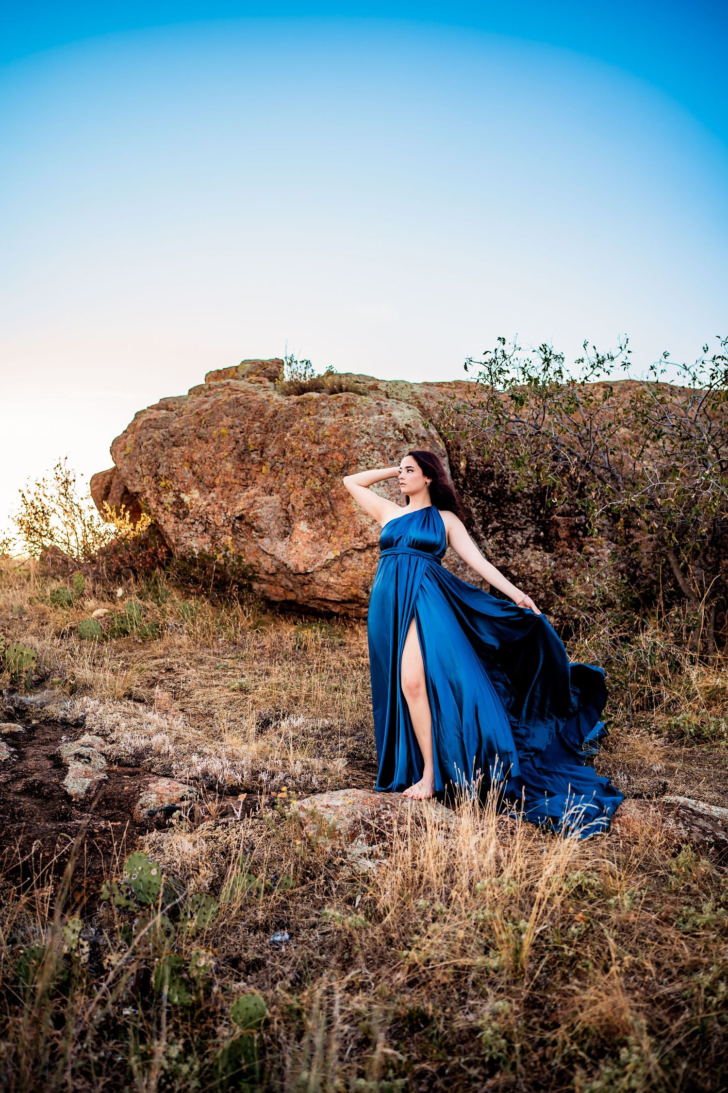 A figure in a flowing blue dress poses gracefully against rocky desert terrain at sunset.