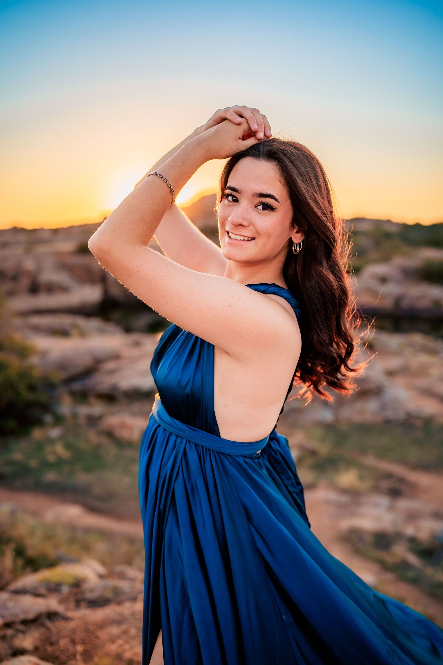 A flowing navy blue dress catches the golden light during sunset in a desert landscape with rocky terrain.