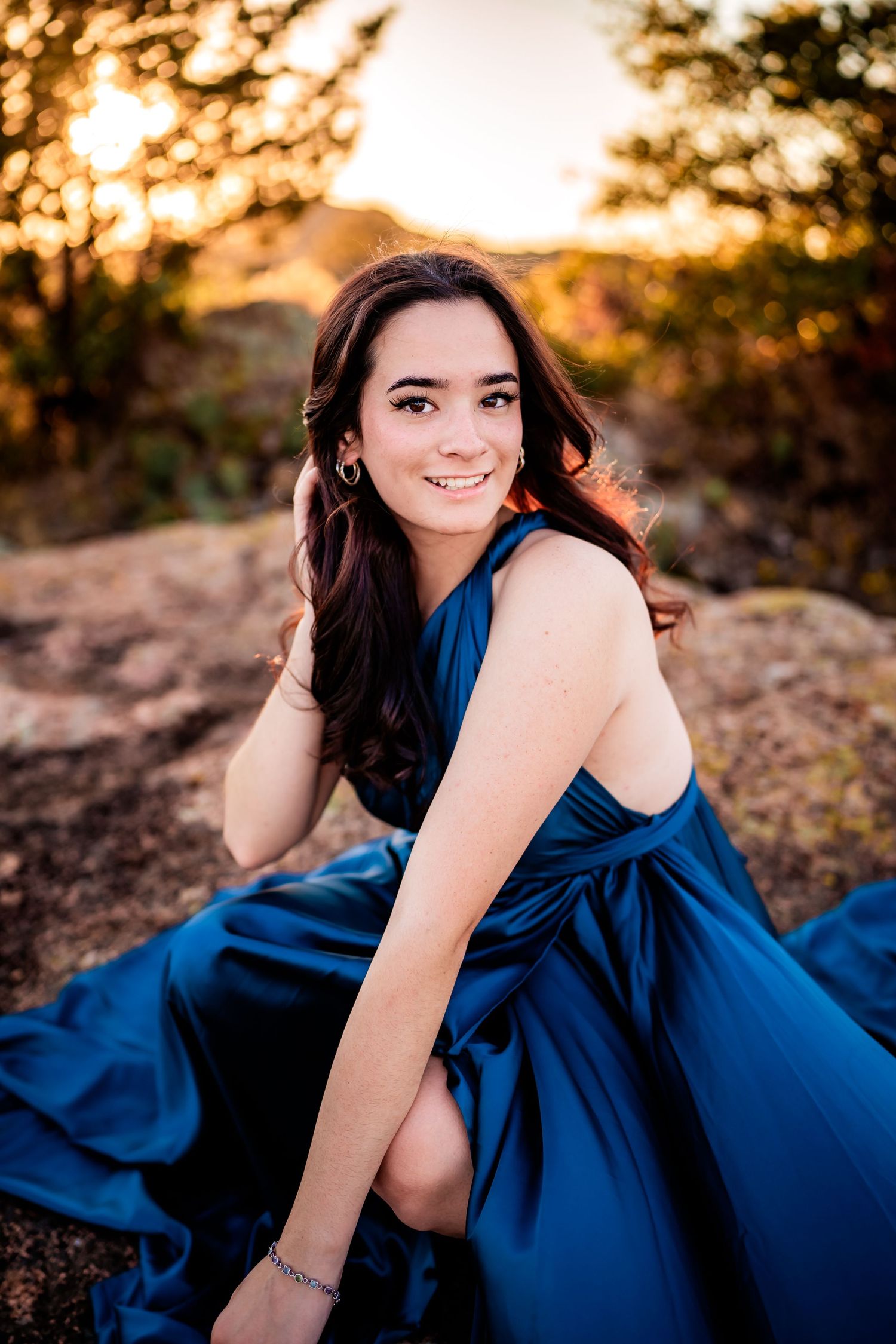 A person in a flowing navy blue dress poses outdoors during golden hour sunset in a rocky natural setting.