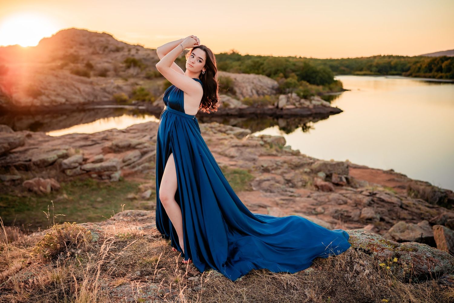 A flowing blue dress billows in the wind against a sunset backdrop at a rocky lakeside location.