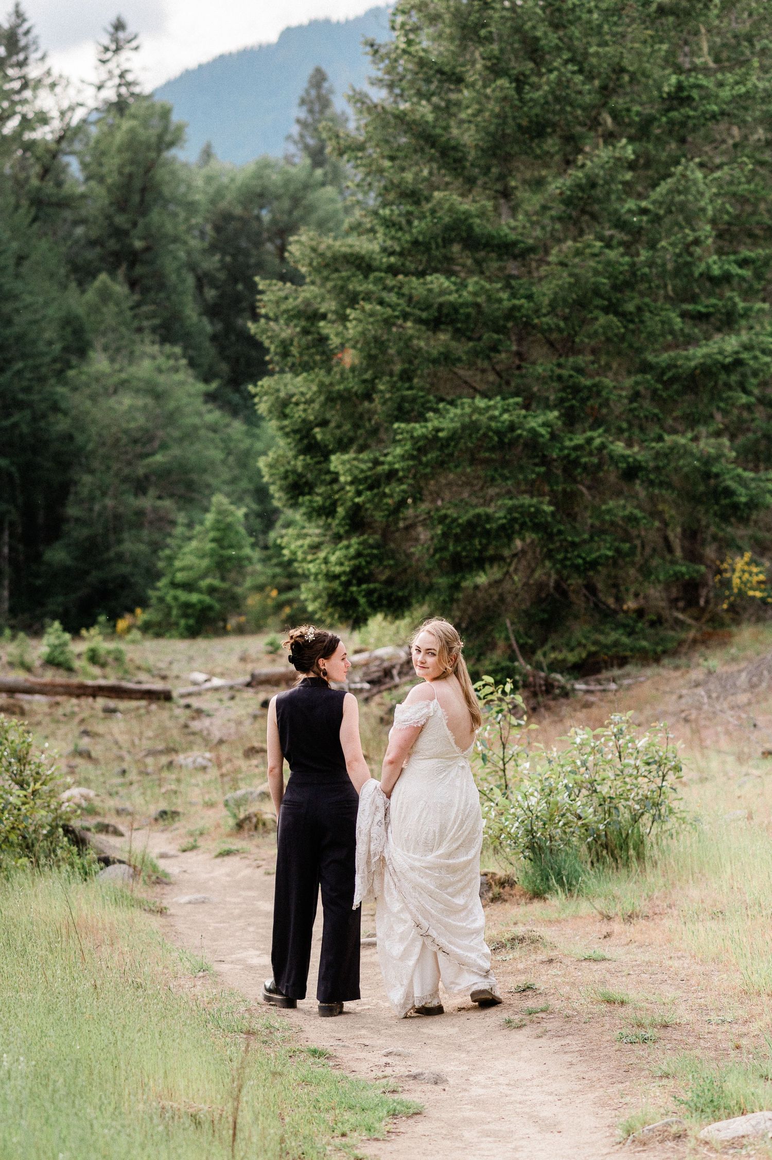 Two brides walk along a dirt trail surrounded by evergreen trees and mountain scenery.