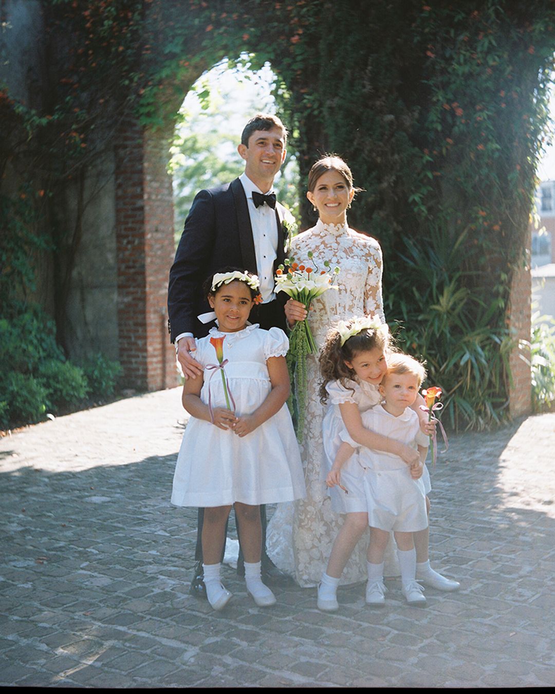Wedding party in white and floral dresses posed on brick pathway in garden setting.