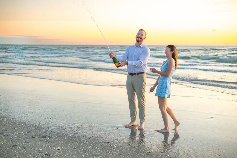 Couple celebrating on beach at sunset, man popping champagne bottle while woman in blue dress walks beside him Lido beach florida