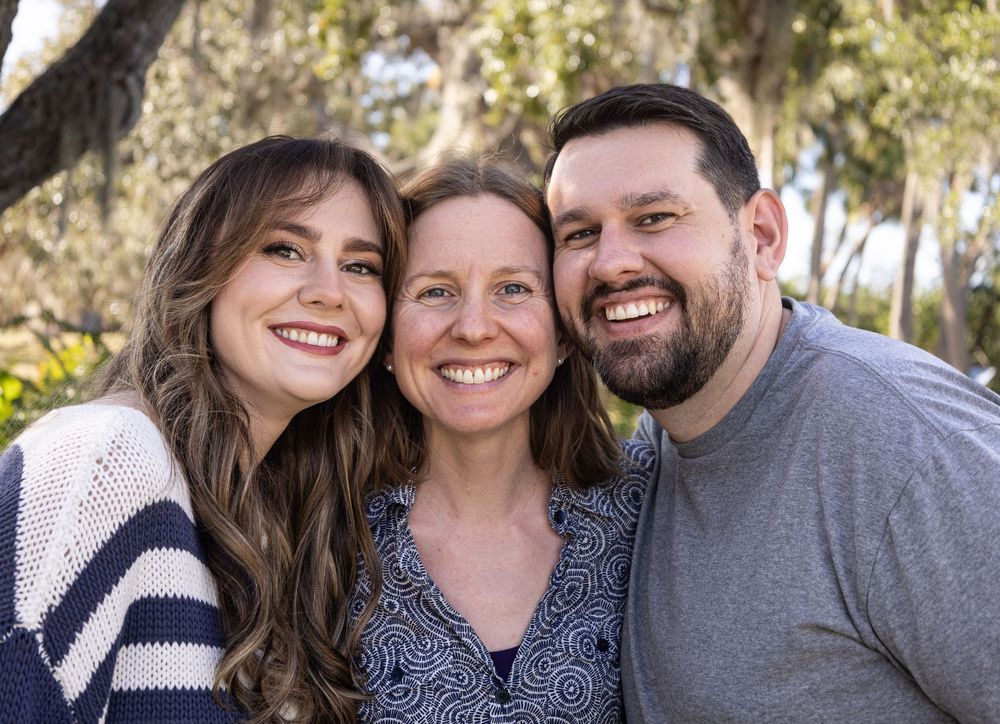 Three smiling friends pose together outdoors among trees in a warm, cheerful group portrait photo  In Venice Florida