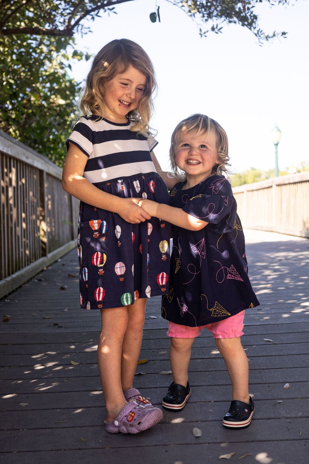 Two young girls in navy dresses smile and hold hands on a wooden boardwalk outdoors In Venice Florida