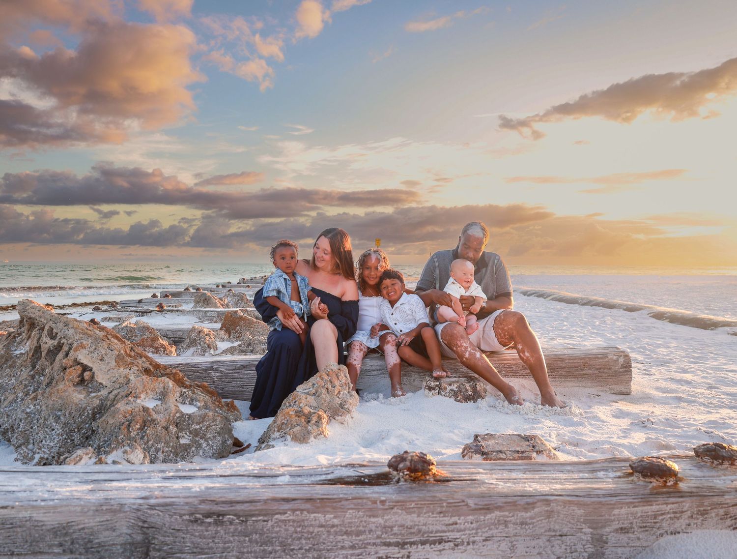 Family group portrait on rocky beach at sunset with ocean waves, warm golden light and colorful sky.