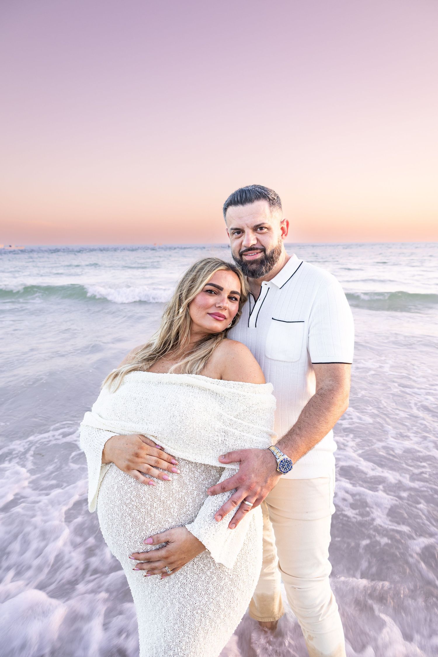 Couple embracing on beach at sunset, pregnant woman in white dress with partner in white shirt